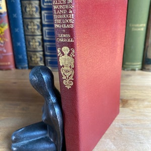 May include: A red hardback book with gold lettering titled "Alice in Wonderland & Through the Looking Glass" by Lewis Carroll. The book is on a wooden surface and is being held up by a black metal bookend shaped like a woman.