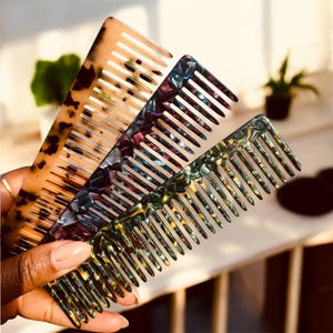 May include: Three decorative combs in a variety of colors are displayed. The first comb has a tortoise shell pattern, the second has a marbled purple and blue design, and the third has a marbled green and yellow design. The combs are held in a hand.