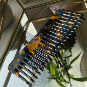 May include: A wide-toothed comb with a marbled design in shades of gold, navy blue, and black. The comb is made of a glossy material and reflects light. The comb is lying on a reflective surface.