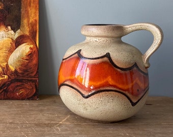 May include: A beige ceramic pitcher with a rounded body and a handle. The pitcher features a bold orange and black zigzag design around its midsection. The pitcher is set on a wooden surface, with a painting in the background.