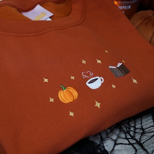 May include: A rust-coloured sweatshirt with an embroidered design of a pumpkin, a steaming cup of coffee, and a closed book. The design is surrounded by small gold stars.