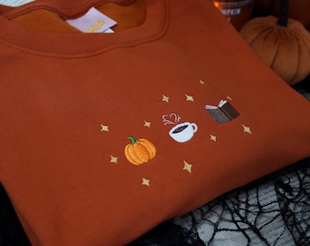 May include: A rust-colored sweatshirt with an embroidered design of a pumpkin, a steaming cup of coffee, and a closed book. The design is surrounded by small gold stars.