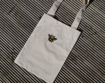 May include: A white canvas tote bag with a yellow and black embroidered bee design.
