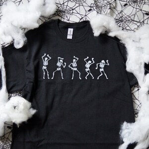 May include: Black t-shirt with a white skeleton dance design. The graphic shows five skeletons in different dancing poses. The shirt is made of a soft material, suitable for Halloween or a themed event.