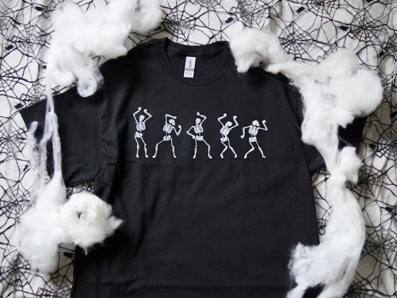 May include: Black t-shirt with a white skeleton dance design. The graphic shows five skeletons in different dancing poses. The shirt is made of a soft material, suitable for Halloween or a themed event.