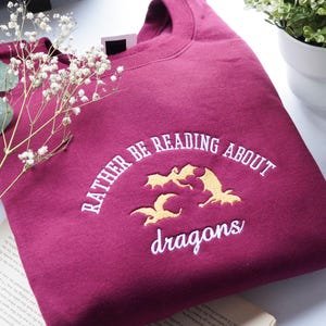May include: A maroon sweatshirt with the embroidered phrase "RATHER BE READING ABOUT dragons" in white and gold. The design features three gold dragon silhouettes. The sweatshirt is folded on top of an open book.