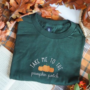 May include: A dark green t-shirt featuring an embroidered design of three orange pumpkins centred on the chest. Above the pumpkins, the text reads "TAKE ME TO THE" in white, and below the pumpkins, the text reads "pumpkin patch" in white.