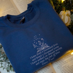 May include: A blue sweatshirt with a white embroidered design featuring a book with flowers and stars. The text reads "I have lived a thousand lives and loved a thousand loves I have walked on distant worlds and seen the end of time because I read."