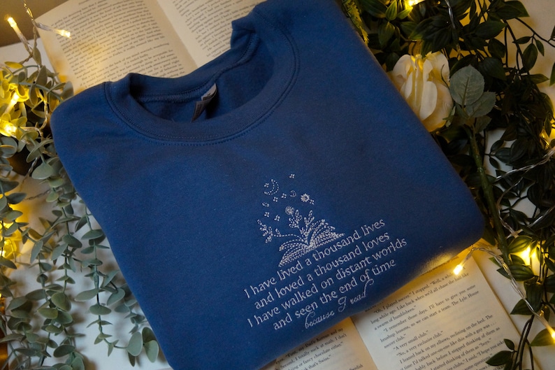 May include: A blue sweatshirt with a white embroidered design featuring a book with flowers and stars. The text reads "I have lived a thousand lives and loved a thousand loves I have walked on distant worlds and seen the end of time because I read."