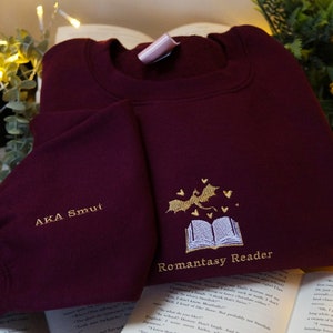 May include: Burgundy sweatshirt with gold embroidery of a book, a dragon, and hearts. The text "Romanstasy Reader" and "AKA Smut" are embroidered on the sweatshirt.