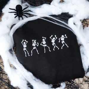 May include: Black tote bag featuring a white dancing skeleton design. The bag is decorated with white spiderwebs, a black spider, and a bat, creating a Halloween theme.