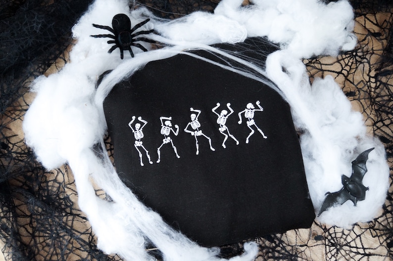 May include: Black tote bag featuring a white dancing skeleton design. The bag is decorated with white spiderwebs, a black spider, and a bat, creating a Halloween theme.