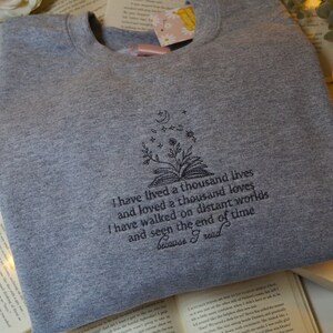 May include: Gray sweatshirt with a black embroidered design of a book with flowers and stars. The text reads "I have lived a thousand lives and loved a thousand loves. I have walked on distant worlds and seen the end of time because I read."