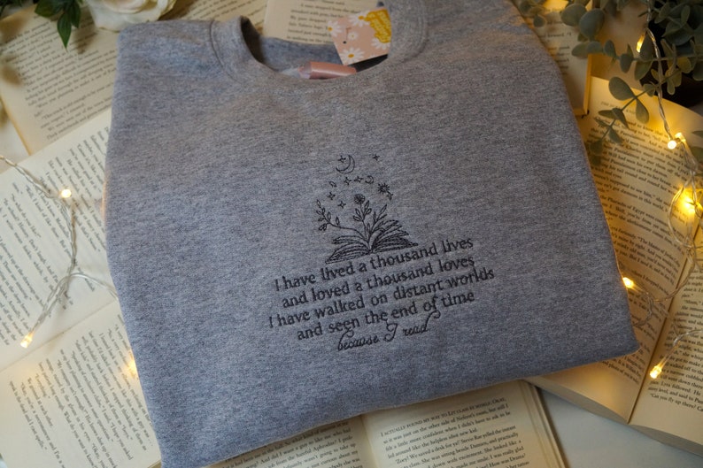 May include: Gray sweatshirt with a black embroidered design of a book with flowers and stars. The text reads "I have lived a thousand lives and loved a thousand loves. I have walked on distant worlds and seen the end of time because I read."