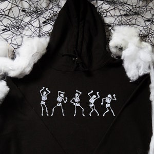 May include: Black hooded sweatshirt with a white graphic of dancing skeletons. The sweatshirt has a drawstring hood and long sleeves. The design is centered on the front. The background includes white cotton and a black spiderweb.