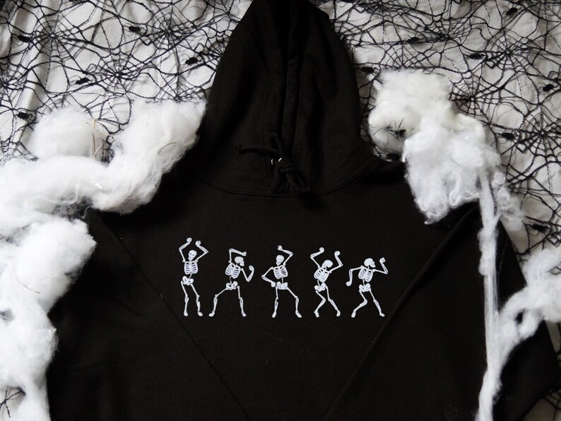 May include: Black hooded sweatshirt with a white graphic of dancing skeletons. The sweatshirt has a drawstring hood and long sleeves. The design is centered on the front. The background includes white cotton and a black spiderweb.
