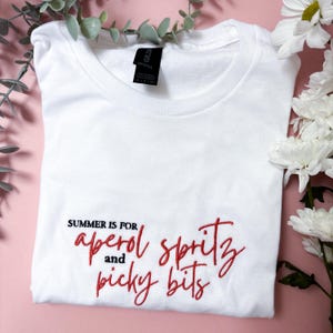 May include: White t-shirt with the embroidered text "SUMMER IS FOR aperol spritz and picky bits" in red script. The shirt is folded and displayed on a pink surface, with white flowers and green leaves in the background.