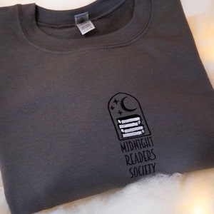 May include: A dark grey sweatshirt with a white embroidered design of a crescent moon, stars, and stacked books. The text "Midnight Readers Society" is embroidered below the design.
