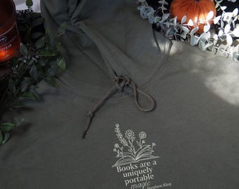 May include: A dark green hooded sweatshirt with a white graphic print that reads "Books are a uniquely portable magic - Stephen King". The graphic also includes a stylized image of an open book with flowers growing from it.