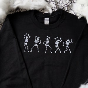 May include: Black crew neck sweatshirt with a white graphic of dancing skeletons. The skeletons are depicted in a line, each in a different dance pose. Ideal for Halloween or themed events.