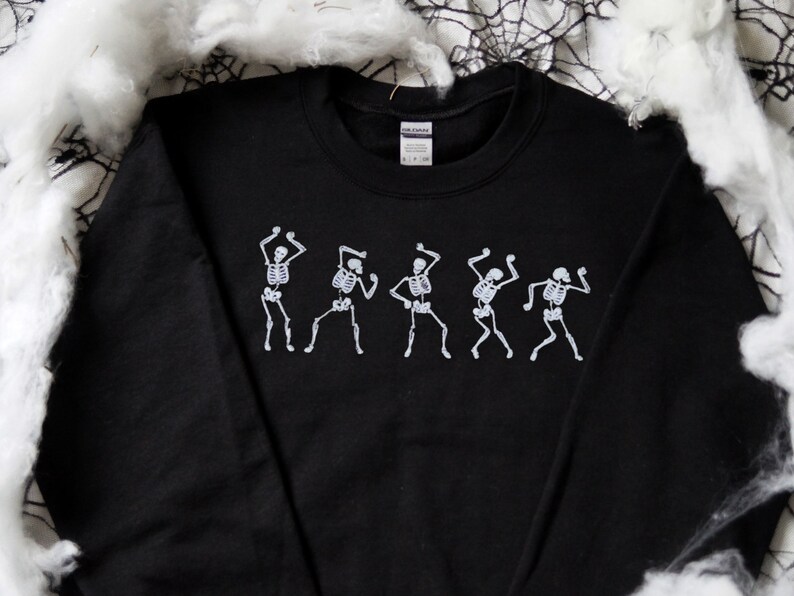 May include: Black crew neck sweatshirt with a white graphic of dancing skeletons. The skeletons are depicted in a line, each in a different dance pose. Ideal for Halloween or themed events.