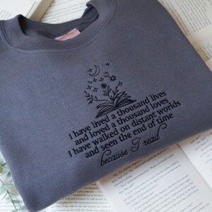 May include: A dark gray sweatshirt with an embroidered design of a book with a flower growing out of it and stars above. The text reads "I have lived a thousand lives and loved a thousand loves I have walked on distant worlds and seen the end of time because I read."