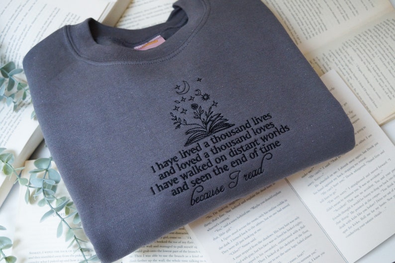 May include: A dark gray sweatshirt with an embroidered design of a book with a flower growing out of it and stars above. The text reads "I have lived a thousand lives and loved a thousand loves I have walked on distant worlds and seen the end of time because I read."