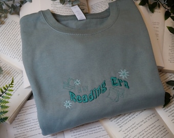 May include: A sage green sweatshirt with the text "It's My Reading Era" embroidered in teal green with stylized flowers and books.
