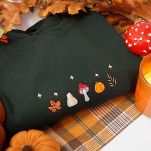 May include: A dark forest green crewneck sweatshirt featuring small, detailed embroidery across the chest. The design includes a vibrant orange maple leaf, a cream-colored gourd, a red and white mushroom, an orange pumpkin, and a brown leaf, all accented by tiny gold embroidered stars.