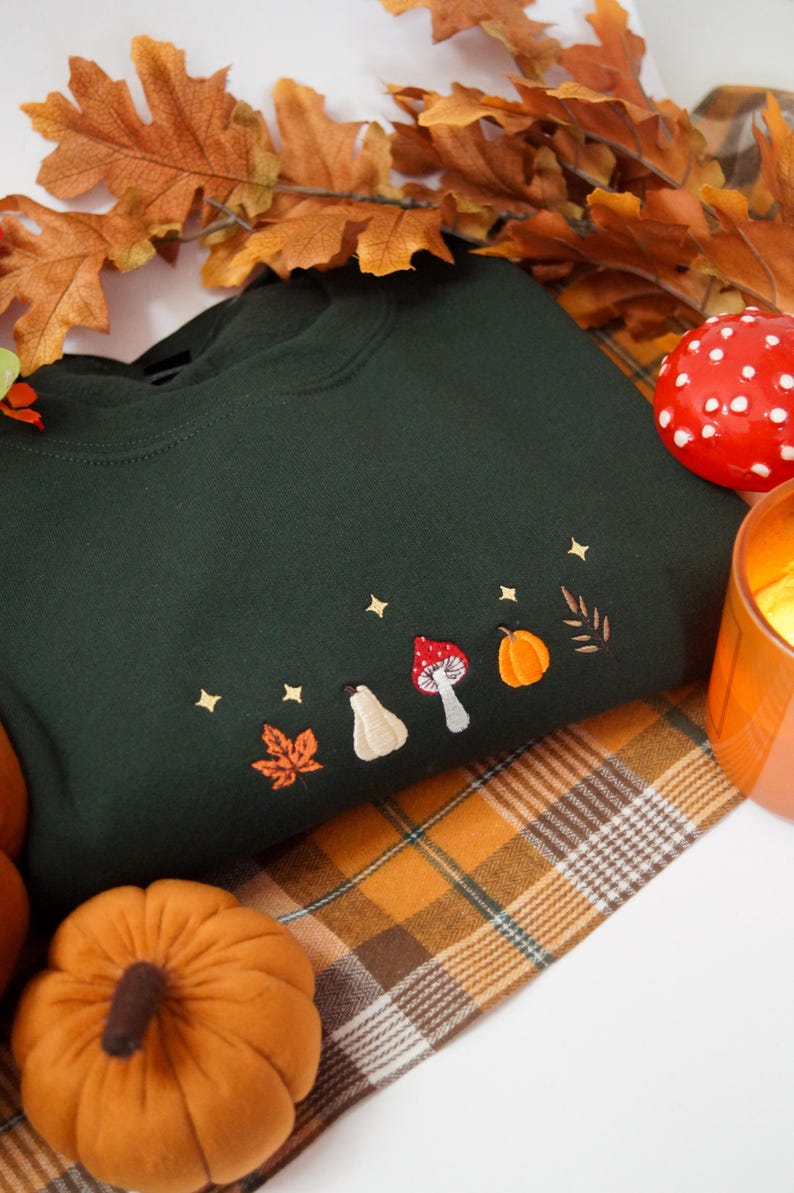 May include: A dark forest green crewneck sweatshirt featuring small, detailed embroidery across the chest. The design includes a vibrant orange maple leaf, a cream-colored gourd, a red and white mushroom, an orange pumpkin, and a brown leaf, all accented by tiny gold embroidered stars.