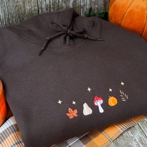 May include: A dark brown hoodie with an embroidered autumn design. The design includes a maple leaf, pear, mushroom, pumpkin, and stars. The hoodie is displayed on a plaid blanket with pumpkins and a candle in the background.