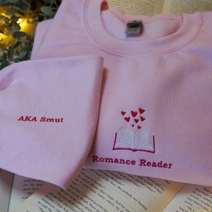 May include: Pink sweatshirt with the text "Romance Reader" and a book with hearts coming out of it. The text "AKA Smut" is embroidered on the sleeve.