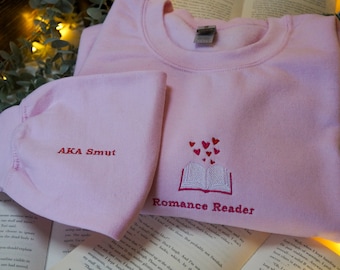 May include: Pink sweatshirt with the text "Romance Reader" and a book with hearts coming out of it. The text "AKA Smut" is embroidered on the sleeve.