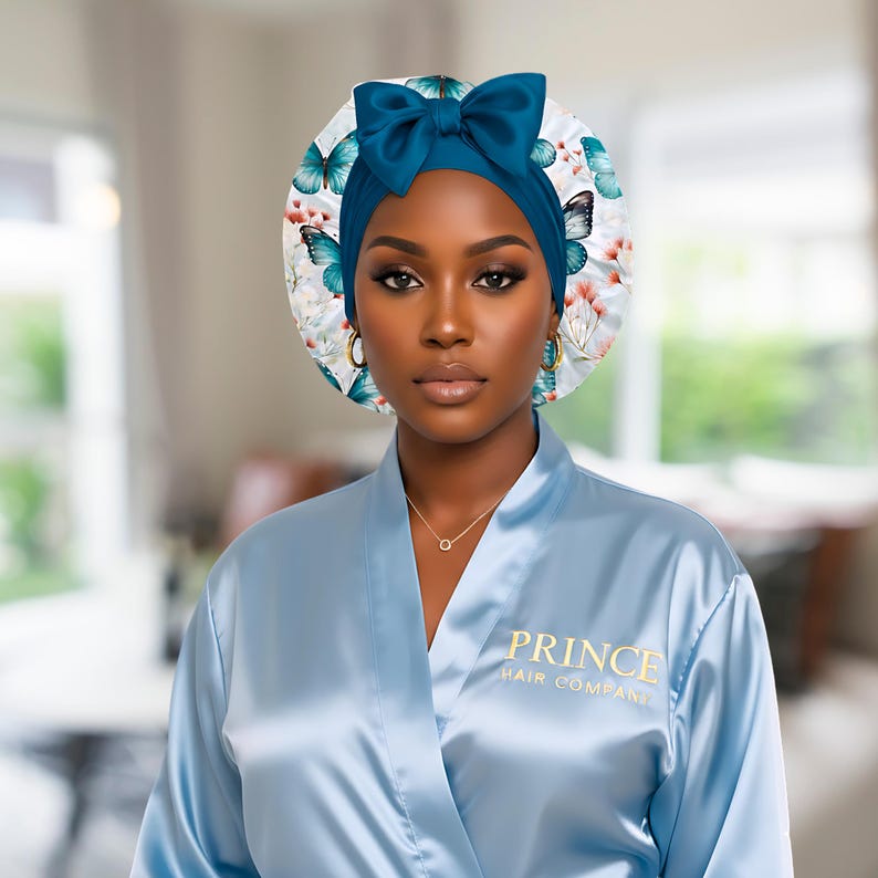 A woman wearing a light blue satin robe with “Prince Hair Company” in gold lettering poses confidently against a blurred background. She wears a blue butterfly-print charmeuse satin bonnet with a matching blue stretch tie and bow, showcasing a polish