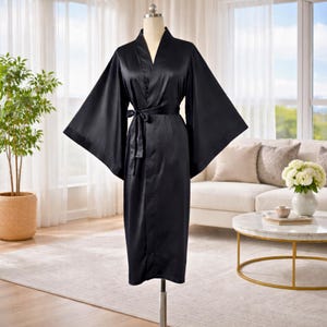 May include: A solid black satin robe featuring wide, kimono-style sleeves, a V-neckline, and a matching fabric tie belt at the waist. The garment is displayed on a mannequin and has a smooth, lustrous finish that reflects light across the fabric.