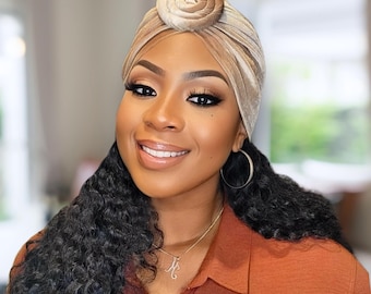 May include: A woman with dark curly hair is wearing a light brown velvet turban with a rose-shaped knot on top. She is wearing a brown shirt and a silver necklace with a letter pendant.