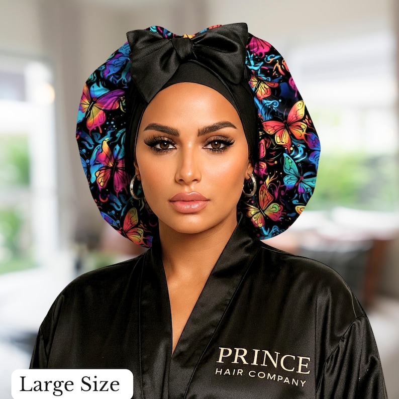 A woman wearing a colorful butterfly-patterned satin hair bonnet and a black satin robe embroidered with “Prince Hair Company” in gold lettering. The soft indoor lighting highlights the bonnet’s silky texture and the brand’s luxury aesthetic. This ha