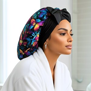 A woman wearing a colorful butterfly-patterned headscarf with a black bow, sitting in a bright modern bathroom. The soft lighting highlights the scarf’s silky texture and vibrant design. This handmade satin head wrap is perfect for protecting natural
