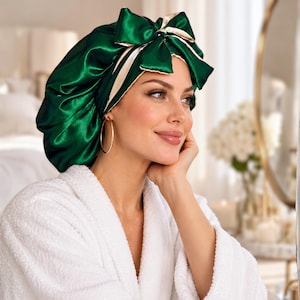 May include: A shiny emerald green satin sleep bonnet featuring a large decorative bow on the front. The interior lining of the bonnet is a contrasting cream color, visible along the edges and the bow. The fabric has a smooth, reflective finish.