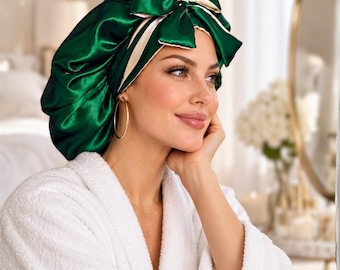 May include: A shiny emerald green satin sleep bonnet featuring a large decorative bow on the front. The interior lining of the bonnet is a contrasting cream color, visible along the edges and the bow. The fabric has a smooth, reflective finish.