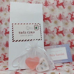 May include: A white gift bag with a red and white label that says "Santa Claus north pole". Inside the bag is a white organza bag with two heart-shaped soaps. One soap is pink and the other is white with dried flowers. A business card for "Bea Savon" is visible in the background.
