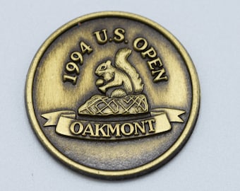 Puede incluir: Una moneda de metal de color dorado con el texto "1994 U.S. OPEN OAKMONT" y una ardilla sosteniendo una nuez.