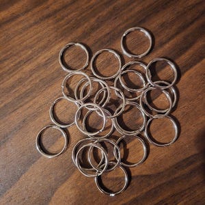 May include: A collection of silver-colored key rings scattered on a dark wood surface. The rings are circular and appear to be made of metal. The key rings are of various sizes and are clustered together.