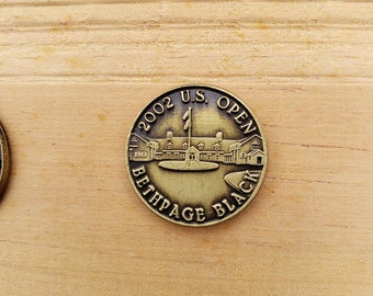 以下が含まれることがあります： 縁に「2002 U.S. OPEN BETHPAGE BLACK」と刻まれた金色の金属製の硬貨。硬貨の中央には、旗が立てられたゴルフコースの小さな画像が描かれている。