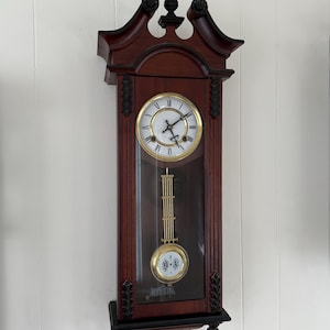 May include: A wooden wall clock with a pendulum. The clock has a dark brown frame with a gold-colored clock face and hands. The pendulum is gold-colored and swings within a glass-covered section. The clock is mounted on a white wall.
