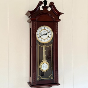 May include: A dark wood wall clock with a white face, black Roman numerals, and ornate hands. The clock features a gold-toned pendulum with the letters R and A, and a glass panel displaying the internal mechanism against a dark background.