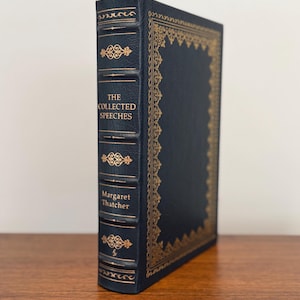 May include: A hardcover book titled "The Collected Speeches" by Margaret Thatcher. The book has a dark blue cover with gold decorative accents and lettering. The book is standing upright on a wooden surface.