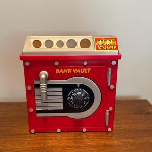 May include: A red "BANK VAULT" coin bank with a combination dial and coin slots. The bank has a silver-colored door handle and a digital display that reads "TOTAL SAVED". The bank is sitting on a wooden surface.