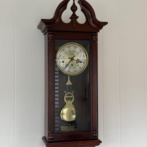May include: A dark wood wall clock with a glass front, featuring a gold-toned circular face with black numerals and hands. A brass-colored pendulum hangs below the clock face within the rectangular wooden frame, which is topped with a decorative arch.