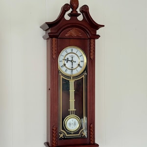 May include: A dark wood wall clock featuring a white circular face with black Roman numerals and ornate black hands. Below the clock face, a glass panel reveals a gold-toned pendulum with a circular center piece displaying the letters H and A.